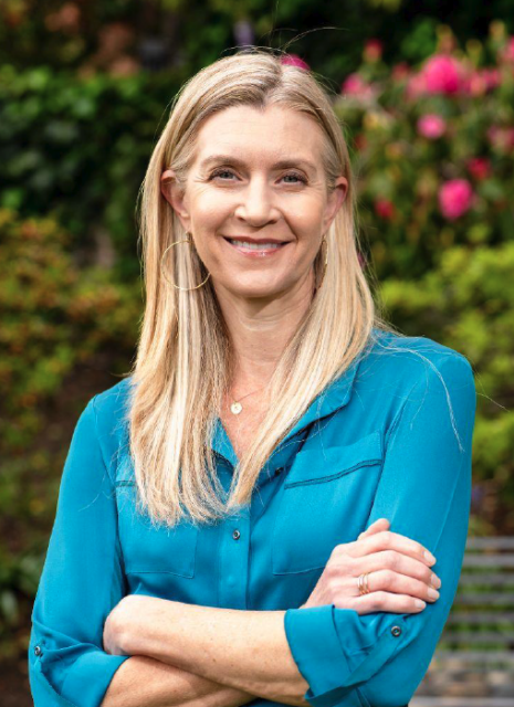 A woman with long blonde hair, wearing a turquoise blouse and hoop earrings, stands outdoors with her arms crossed. Pink flowers and greenery are in the background.